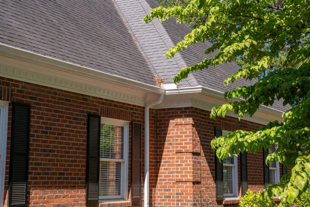 A white gutter and downspout at a brick corner under a tree